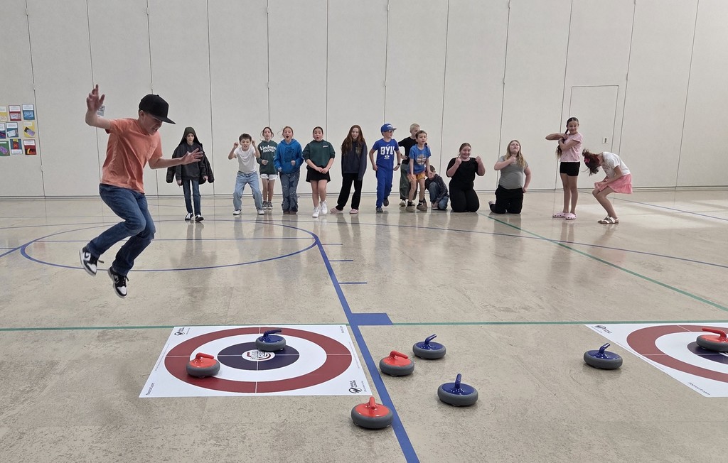 A young boy in an orange t-shirt and black baseball cap mid-air, jumping with excitement on a gymnasium floor. In the background, a line of students watches and cheers. In the foreground, red and blue floor curling stones are positioned on and around a target mat