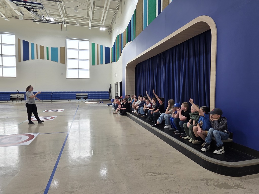 A wide shot of a school gymnasium where an instructor in a grey shirt is gesturing to a large group of students. The students are sitting in a row along the edge of a stage with blue curtains. Several students have their hands raised. Three floor curling target mats are lined up on the left side of the gym floor.