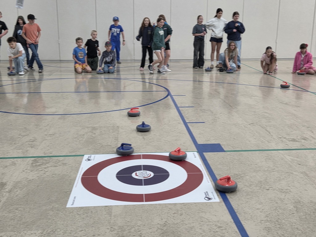 A close-up perspective of red and blue floor curling stones sliding across a gym floor toward a circular target mat. Students are standing in the background, observing the progress of the game.