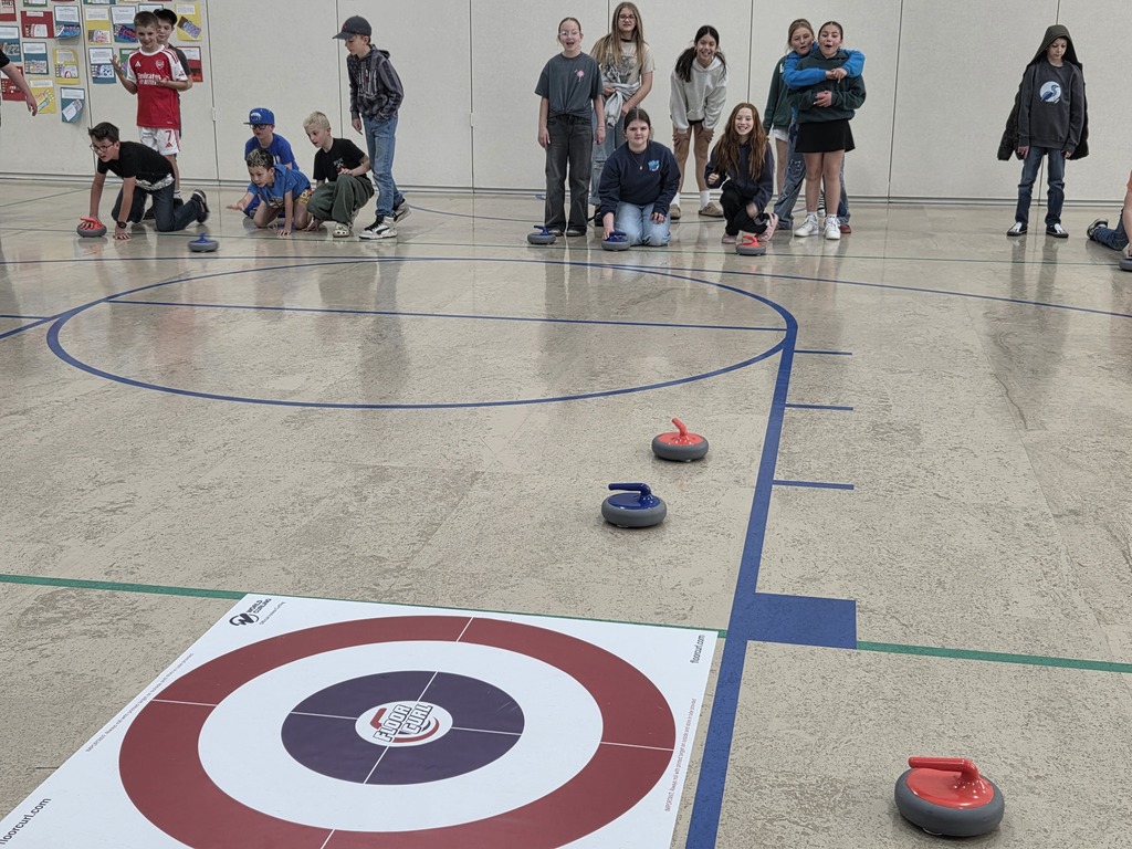Two groups of students face each other across a gymnasium floor during a game of floor curling. Several students are crouched down, ready to slide stones toward a target mat in the foreground.