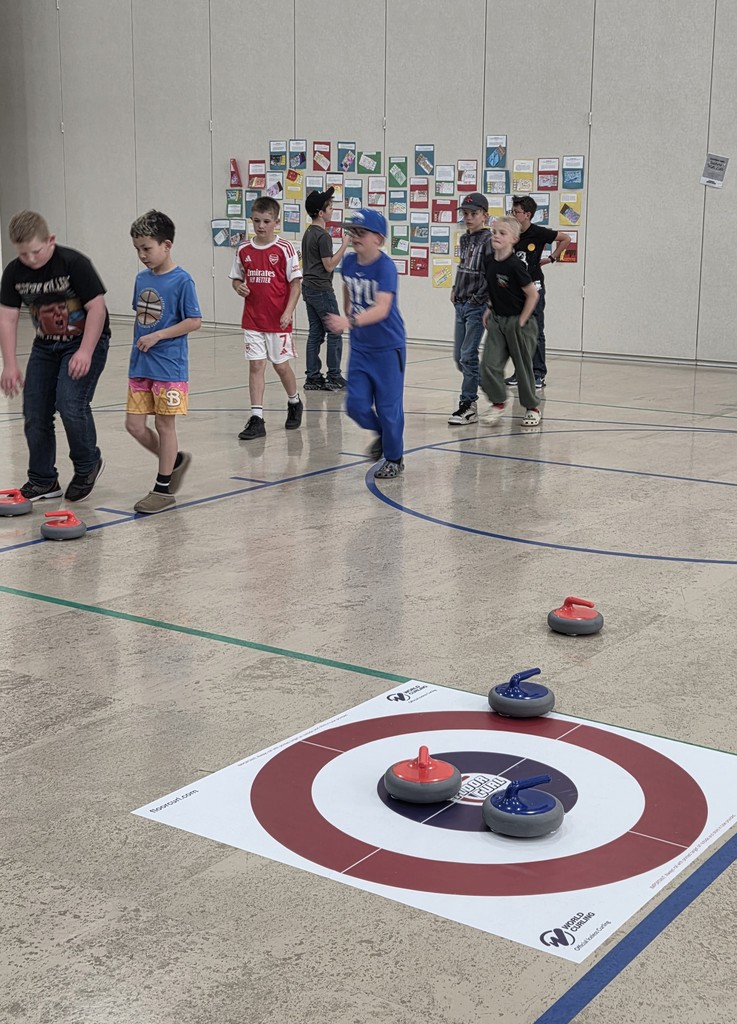 A group of boys walking across a gym floor toward a floor curling target. One boy wears a red Arsenal soccer jersey and another wears a blue BYU shirt. Red and blue curling stones are scattered on the polished floor.
