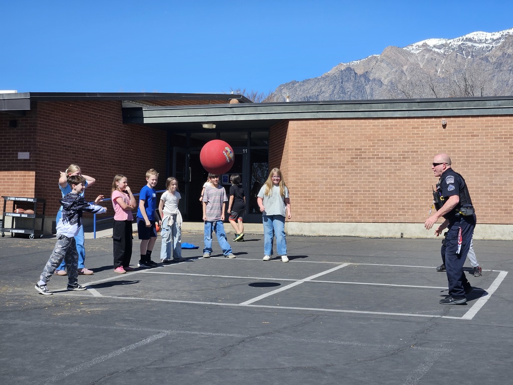 A sunny outdoor playground at Lomond View Elementary with mountains in the background. A group of elementary school students stands around a four-square court, watching a police officer in uniform lean forward to hit a large red playground ball. Everyone is smiling and engaged in the game.