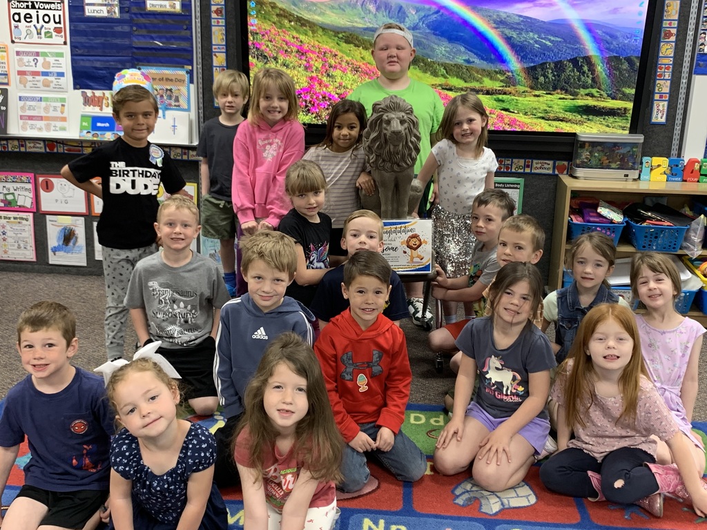 A group photo of Mrs. Larson's joyful kindergarten class gathered together in their classroom, smiling. A large, stone-like lion statue, Sir Fluffington, is the central focus, resting on a cart with a small celebratory sign that reads "Congratulations on having the most Awesome behavior!" behind it. In the background is a large screen displaying a mountain and rainbow scene. The children are posed around the statue, some wearing birthday crowns and special shirts. The room is decorated with colorful educational posters and storage bins.