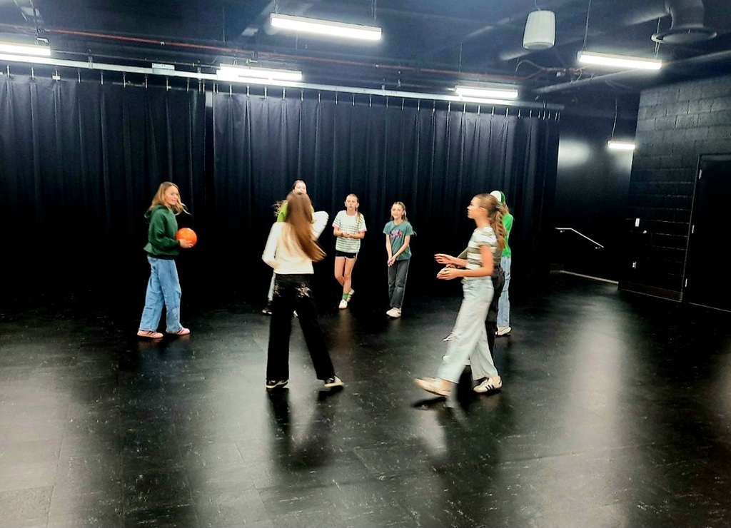 Six young girls standing in a circle on a dark, polished stage or studio floor, surrounded by heavy black curtains. One girl on the left in a dark green hoodie and jeans holds an orange ball. The room is lit by overhead fluorescent strip lights.