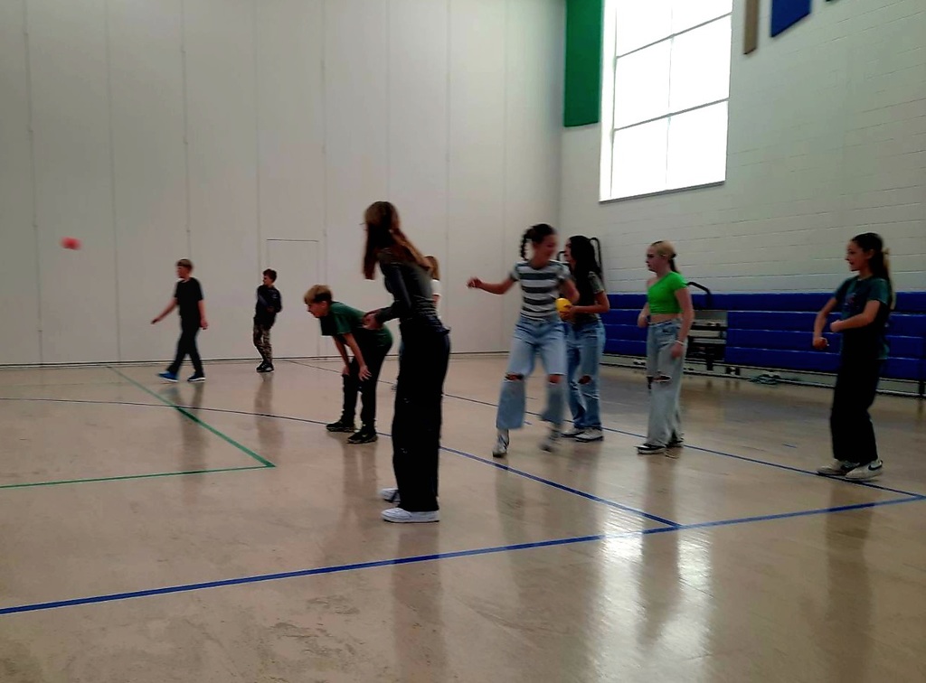 A group of middle-school-aged students playing dodgeball in a bright gymnasium with white walls and blue bleachers. A blurred red ball is in mid-air on the left side of the frame. Students are seen in various athletic poses, including a girl in a striped shirt and light-wash ripped jeans ready to move, and another girl in a bright green top. The gym floor has green and blue court markings.