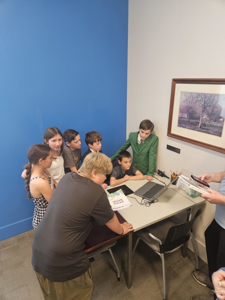 A group of students gathered around a desk in a "CFO" office, focused on a laptop and Zions Bank documents during a business simulation.
