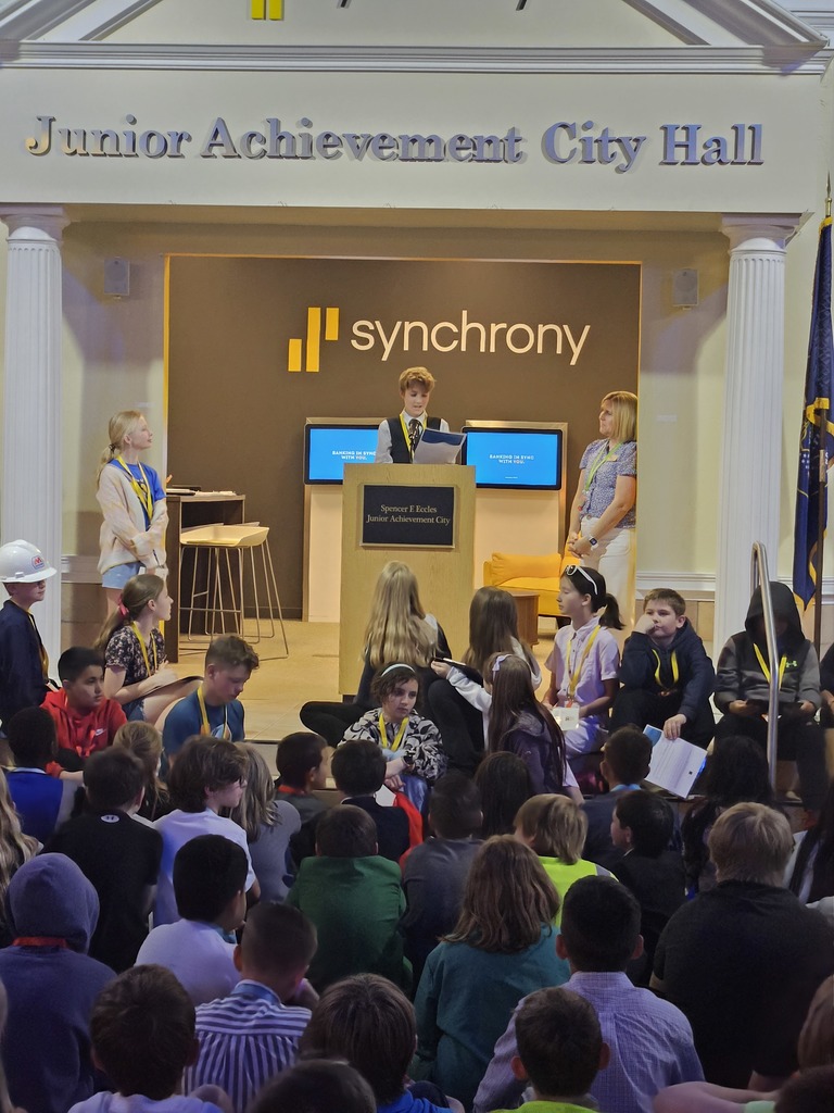 A student stands at a podium giving a speech in front of a seated audience at the Junior Achievement City Hall, sponsored by Synchrony.