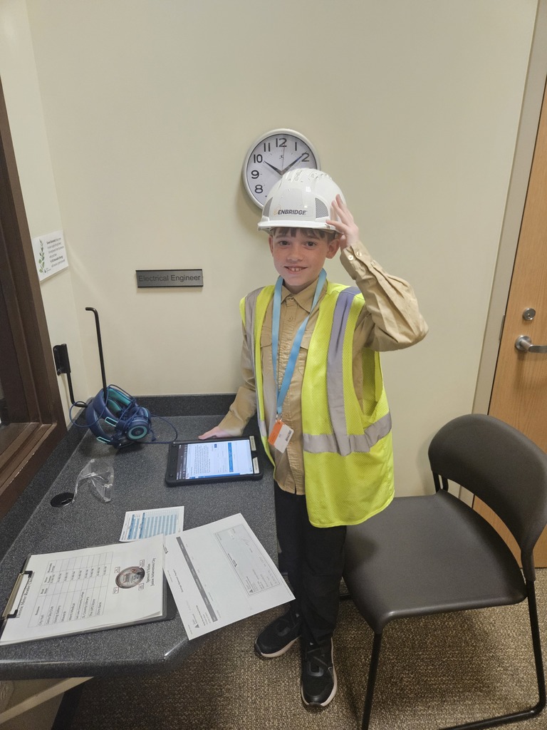 A smiling student dressed as an Electrical Engineer, wearing a neon safety vest and hard hat, standing at a workstation with a tablet and paperwork.