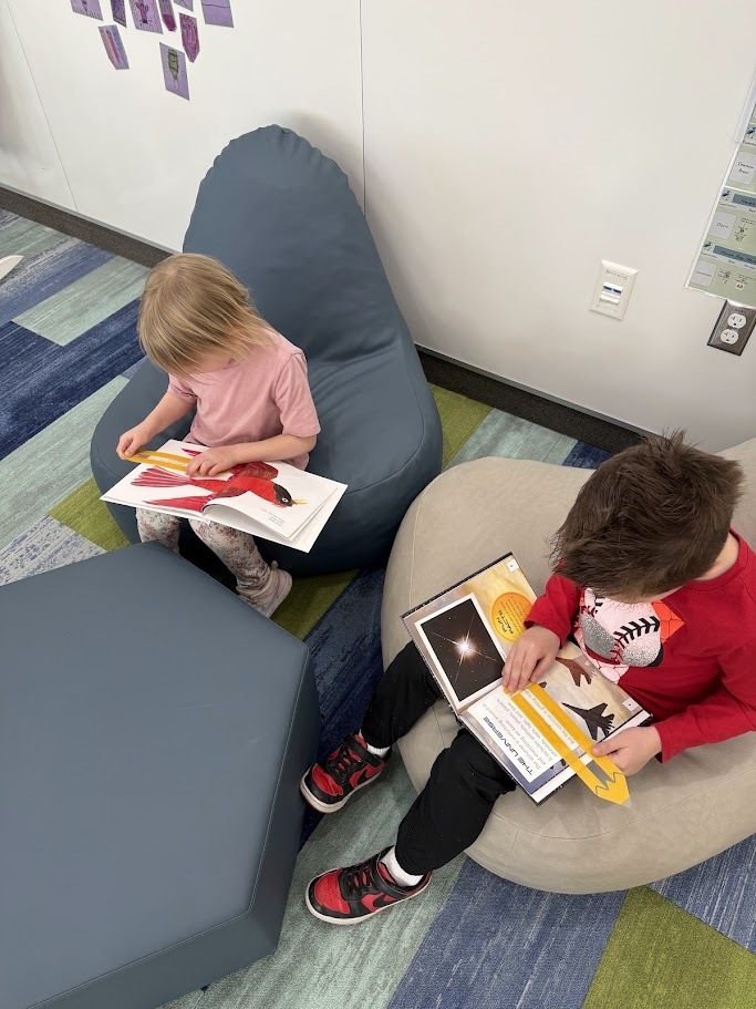 High-angle view of two children sitting in bean bag chairs reading. A girl in a pink shirt looks at a book featuring a large red bird. A boy in a red shirt reads a book titled THE UNIVERSE with an image of a star. Both use yellow, pencil-shaped reading trackers.