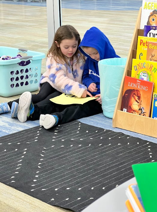 Two young children sit on a blue patterned floor next to a wooden bookshelf. A girl in a floral fleece jacket points to a yellow paper held by a boy in a blue Champion hoodie. Bookshelf includes titles like The Lion King, Unicorn, and Pete the Cat.