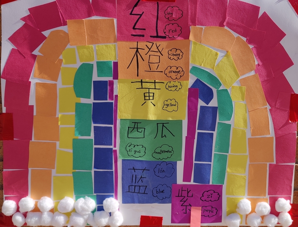 A rainbow arch made from small, mosaic-style pieces of construction paper. The colors move from red on the outermost layer to orange, yellow, green, and blue on the inner layers. There is A vertical stack of colored paper rectangles contains educational text. From top to bottom: Red: "红" (hóng / red) Orange: "橙" (chéng / orange) Yellow: "黄" (huáng / yellow) Green: "西瓜" (xī guā / watermelon) Blue: "蓝" (lán / blue) Purple (offset to the side): "紫" (zǐ / purple).