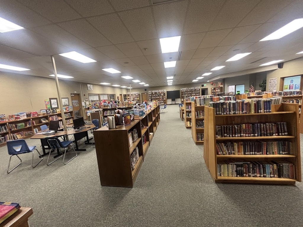 A wide-angle interior view of a school library with carpeted floors and fluorescent lighting. Numerous wooden bookshelves, stocked with books, create long aisles. A study area with tables, multiple computer monitors, and blue chairs is visible in the background, and smaller blue chairs are scattered throughout the space.