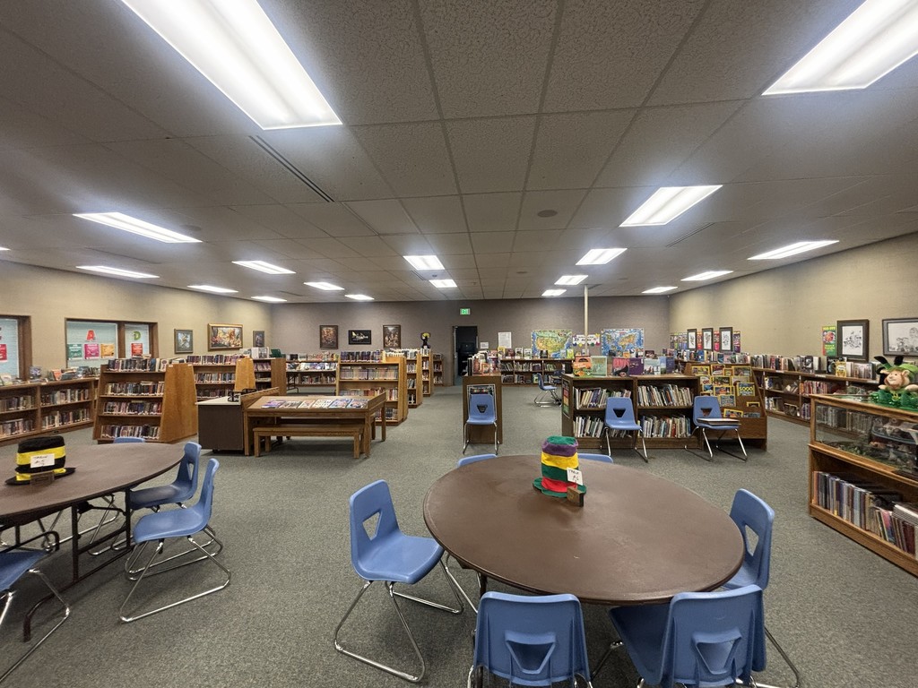 A wide view of the Bates Elementary library featuring two large round wooden tables in the foreground with blue chairs. The spacious room is filled with several rows of wooden bookshelves, a low display table for books, and various educational posters on the neutral-colored walls.