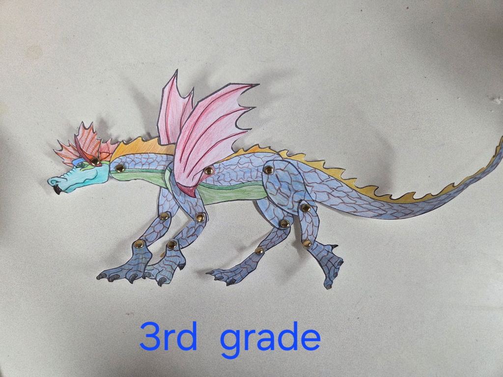 A photo of a 3rd-grade student's articulated paper dragon against a neutral white background. The dragon is made of blue and green textured paper with pink-veined wings. Metal split pin fasteners are visible at the joints of the head, body, wings, and all four legs. Text in blue below the dragon reads "3rd grade."
