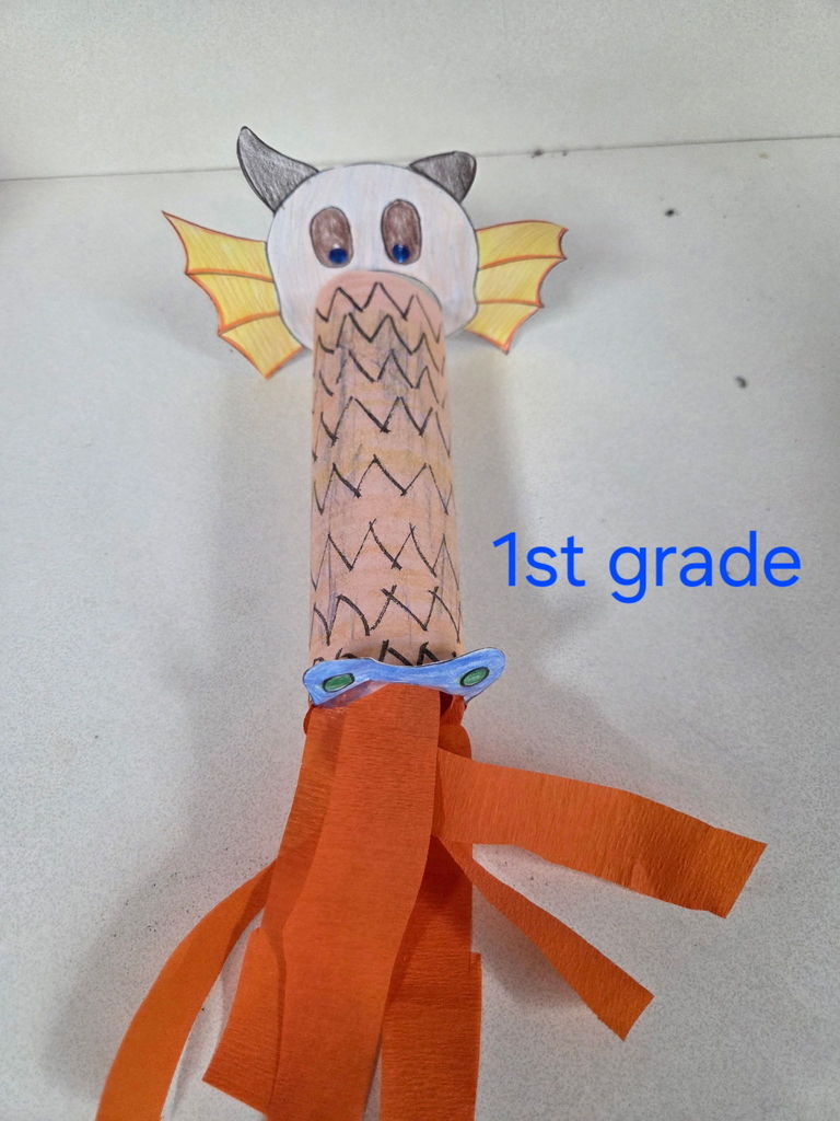 "fire-breathing" dragon crafts from the Bates Elementary 1st-grade class. Each dragon is constructed from a cardboard tube decorated with hand-drawn scales, featuring paper heads with horns, small yellow wings, and blue-jeweled eyes. Long, vibrant strips of orange crepe paper extend from the "mouths" to mimic flames. The top-left panel includes the school's bison mascot in the corner. In the center of the collage, a colorful, rainbow-lettered title reads "1ST GRADE."