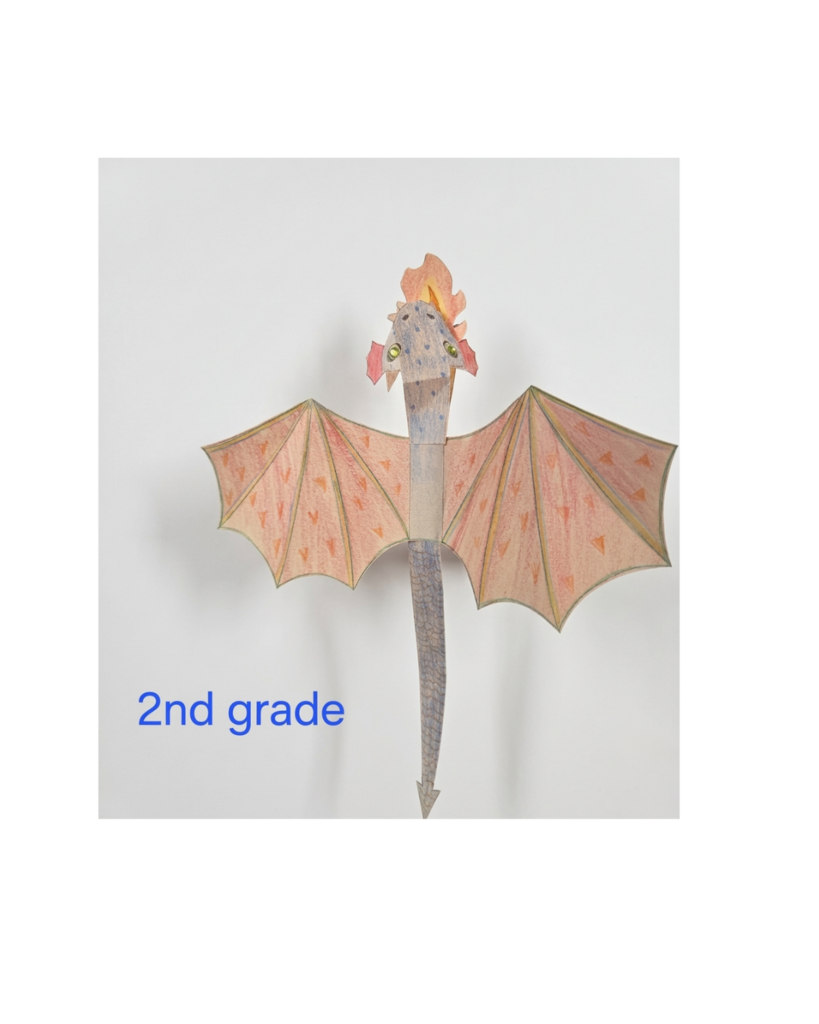 photo of a 2nd-grade paper dragon craft against a plain white background. The dragon has a long, slender blue-grey body with a pointed tail and large, spread-out orange wings decorated with small triangular patterns. The dragon's head features small red ears, yellow-jeweled eyes, and a paper "flame" emerging from its head. In the bottom-left corner, blue text reads "2nd grade."