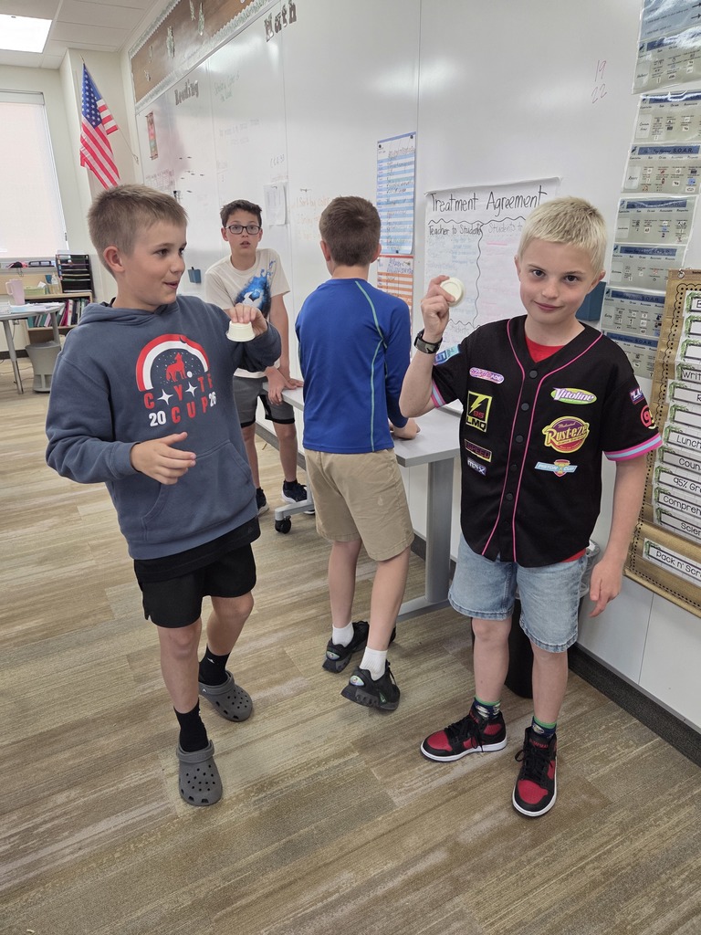 Three boys pose in front of a classroom whiteboard. The boy on the left in a black baseball jersey and the boy in the middle in a grey "COYOTE CUP 2026" hoodie are holding up small white lids. The boy on the right wears a blue and green patterned athletic shirt and khaki shorts. The whiteboard features a "Science" heading, a "Classroom Expectations" chart, and a calendar showing the date "March 26, 2026." Text on the board also mentions "Points Goal 168/175," "Working Towards: Pizza Party," and "Currently Working on: Engagement."