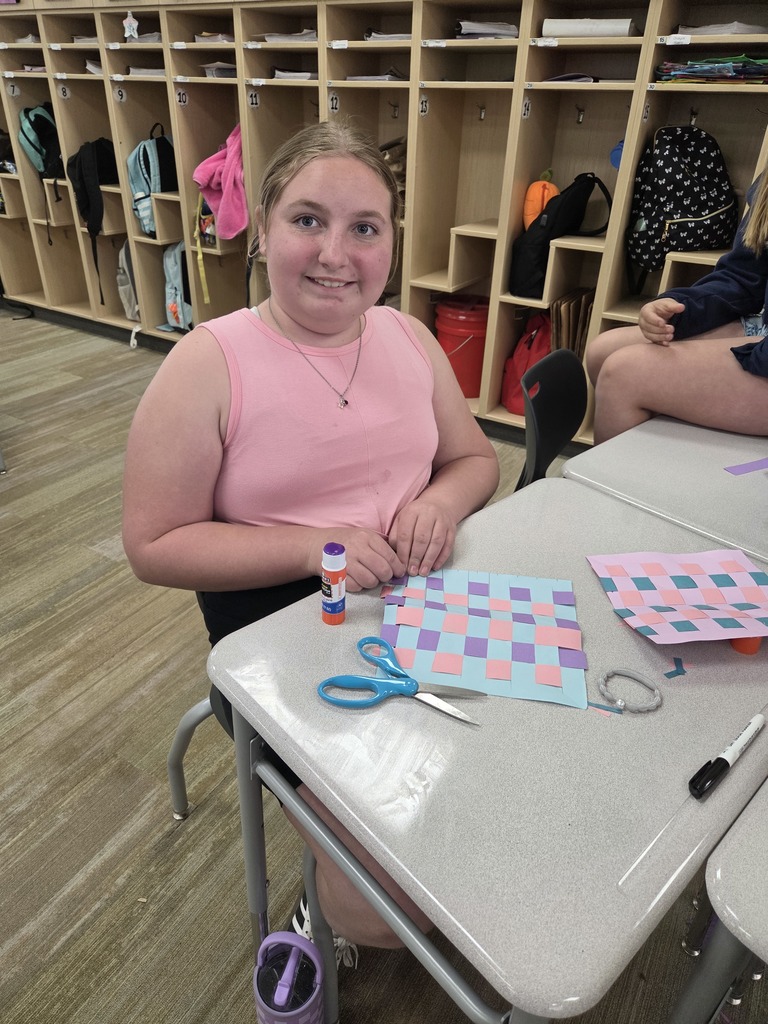 Here are the alternative texts for your images, designed to be descriptive and inclusive of all visible text.  Image 1: Student with Paper Weaving Alt Text: A young female student with light hair and a pink tank top sits at a classroom desk, smiling at the camera. In front of her is a colorful paper-weaving project made of light blue, pink, and purple strips. On the desk are blue scissors, a purple glue stick, a grey bracelet, and a black marker. In the background are wooden classroom cubbies containing backpacks and jackets. The glue stick label reads "ELMER'S Washable School Glue Stick."