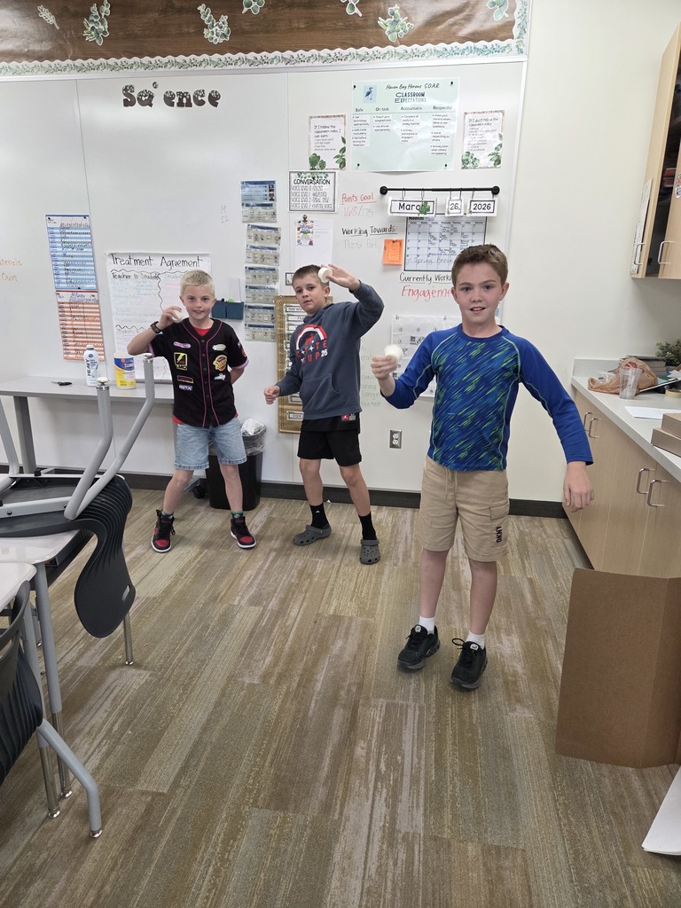 Three young boys stand in a classroom near a whiteboard. Two of the boys are holding up small, white circular containers. One boy on the left wears a grey hoodie with a logo that says "COYOTE CUP 2026." The boy on the right wears a black baseball jersey with several patches including "Vitoline," "95 LMO," "Rust-eze," "OCTANE GAIN," and "Piston Cup." On the whiteboard, a poster titled "Treatment Agreement" lists "Teacher to Students" and "Students to Teacher." Other visible text includes "Reading," "Math," and a daily schedule.