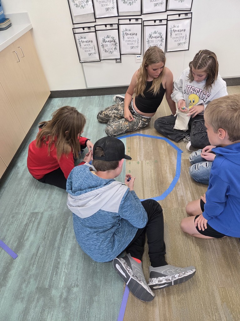 Five students are sitting in a circle on a classroom floor, which is marked with blue tape in an octagonal shape. They are focused on a game involving small green and blue marbles scattered inside the tape. One student holds a small beige drawstring bag. On the wall behind them, several black-bordered frames hold papers that say "Amazing work coming soon!" and "coming soon!" One student wears a grey sweatshirt with the word "JUSTICE" and a Tweety Bird graphic.