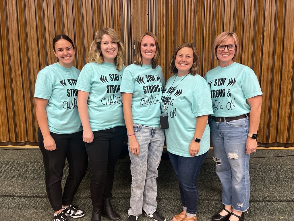 A group photo of five smiling women from the Lomond View Elementary Special Education team. They are standing in a row wearing matching light teal t-shirts with the school’s motivational motto, "STAY STRONG & CHARGE ON," printed in black.