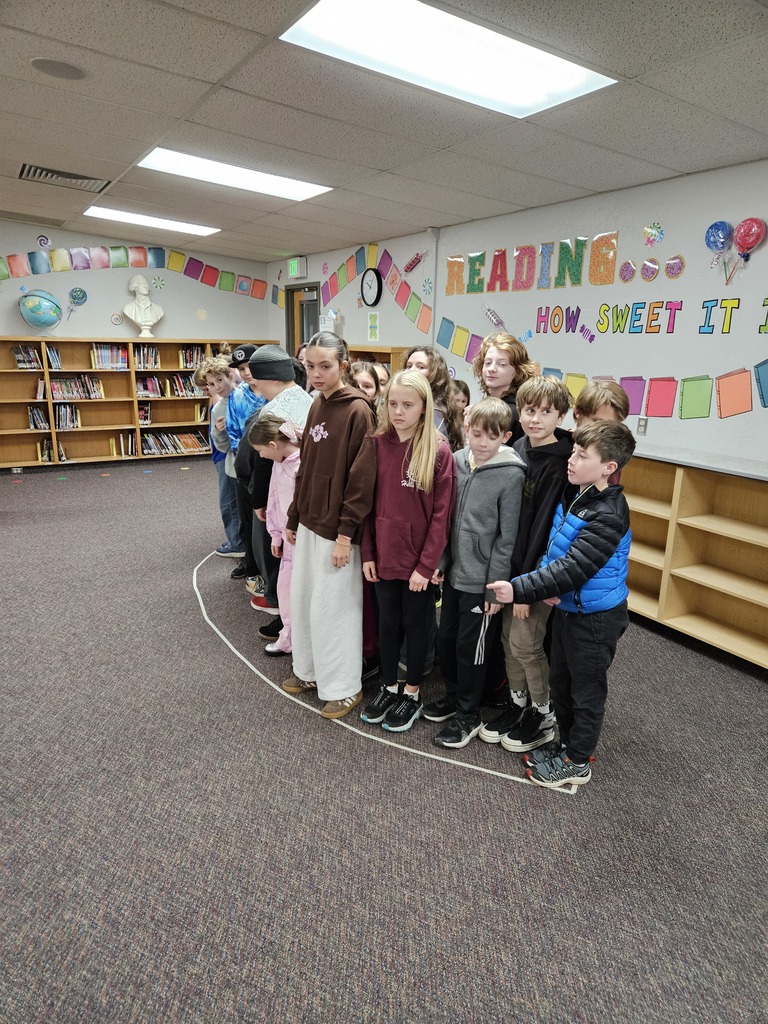 A large group of students standing along a curved white tape line on the library floor. The line represents the scale of a single eye from the Mount Rushmore monument.