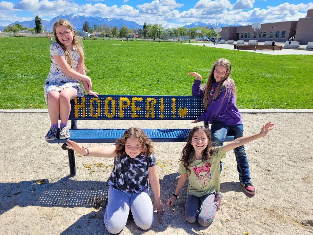 Four girls are posed around a blue metal bench that has "HOOPER!!" written in yellow cups through its mesh backrest. The bench is situated on a sandy area, likely a school playground, with a lush green field and snow-capped mountains in the background.