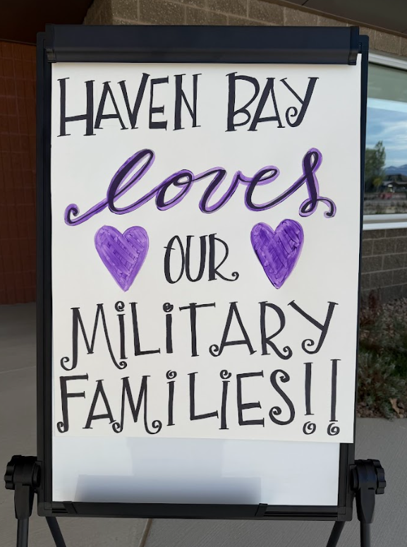 A white sign on an easel featuring hand-drawn text and illustrations. The top line reads "HAVEN BAY" in black stylized letters, followed by the word "loves" in purple cursive. Below are two purple hearts flanking the word "OUR" in black. The bottom reads "MILITARY FAMILIES!!" in large, black decorative block letters.