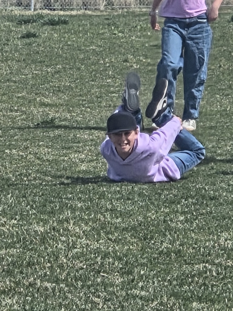 A close-up of a student wearing a black cap and a light purple hoodie, laying on his stomach on a green grass field. He is performing a "seal" or "bow" stretch, reaching back to grab his ankles with his legs bent upward. Another student's legs are visible walking past in the background.