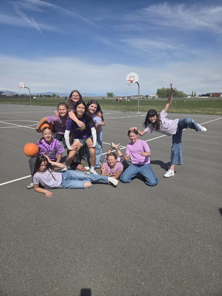 Nine girls pose for a photo on an outdoor basketball court. Some are sitting or reclining on the asphalt while others stand behind them. One girl on the right is balanced on one leg in a star pose. Two basketballs are held by the group on the left. The background shows a field and distant mountains.