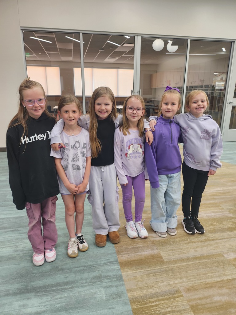 Six young girls stand in a line in a school hallway with glass windows in the background. Most are wearing shades of purple.