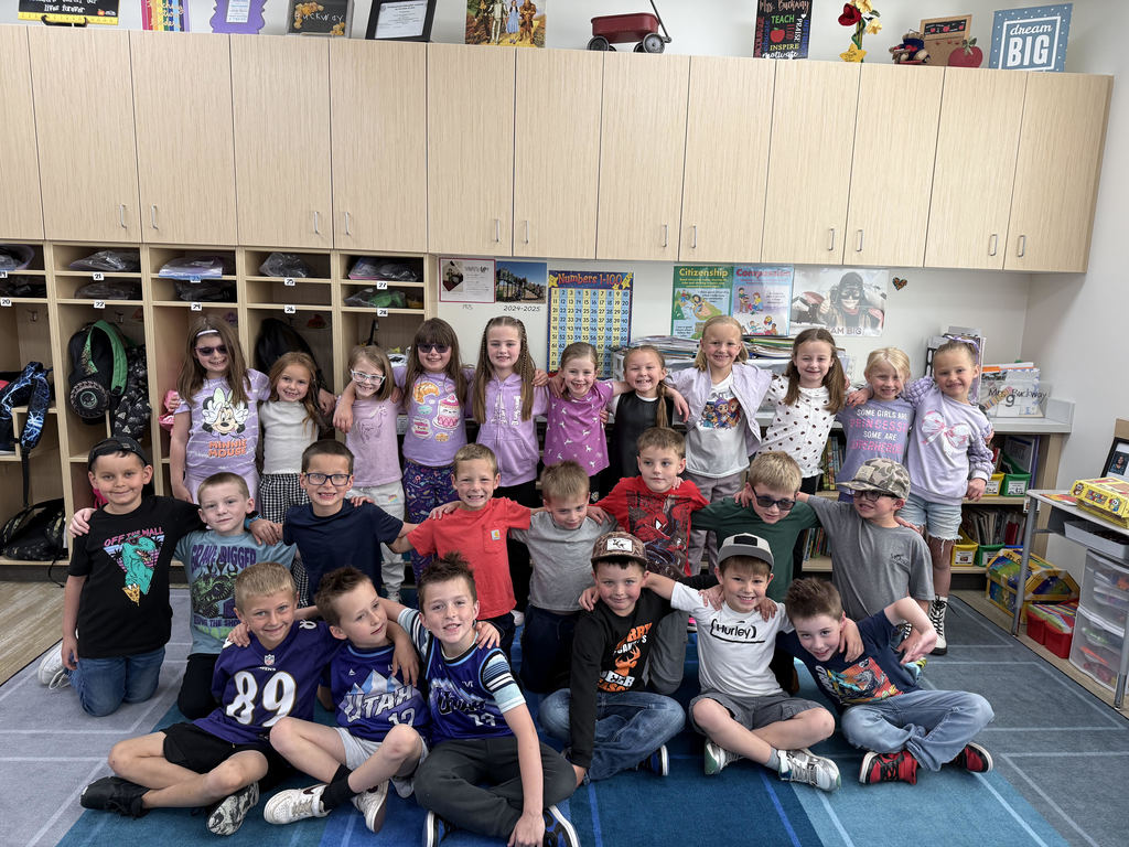 A large group of approximately 25 elementary school students posing in a classroom in front of light wood cabinets. Many students are wearing purple or blue clothing.