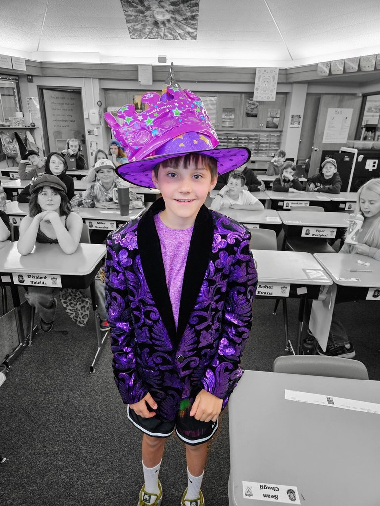 A young male student standing in a classroom wearing an elaborate, sequined purple blazer and a large purple wizard hat topped with a purple paper crown. Other students sit at desks in the background.