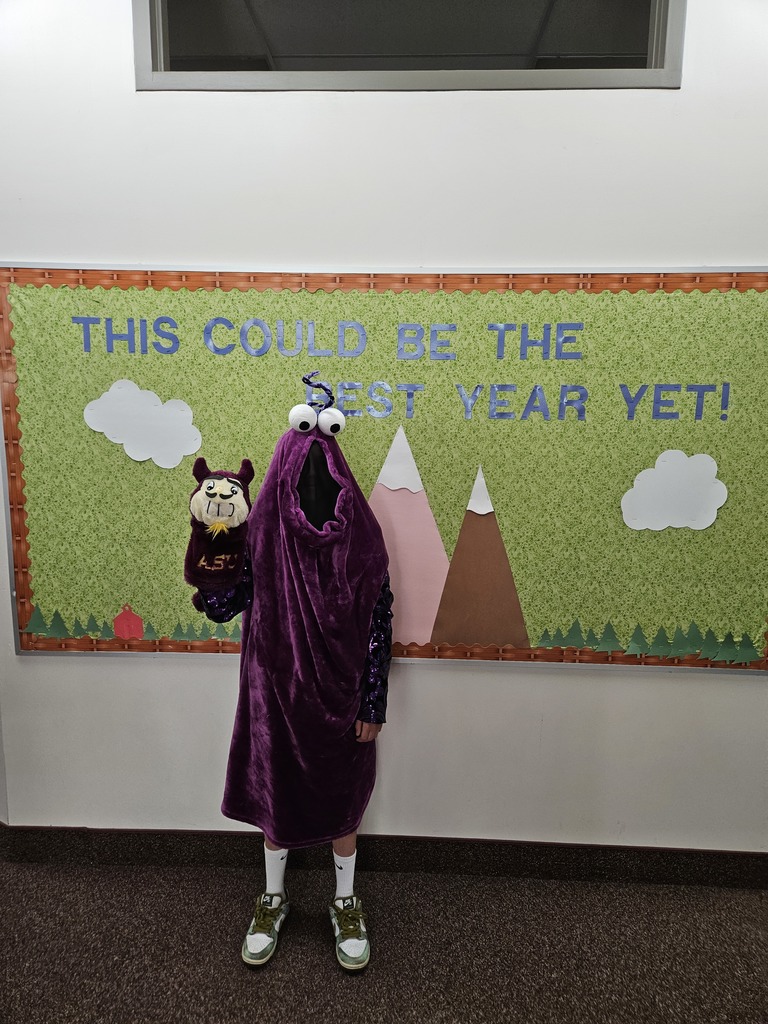 A student completely covered in a fuzzy purple "monster" sack costume with large googly eyes on top, holding a purple puppet. They are standing in front of a school bulletin board .