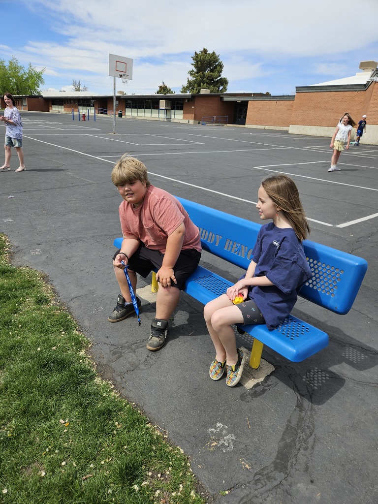 The same young boy is now sitting on the blue "BUDDY BENCH" and looking toward a new friend. A young girl is sitting beside him on the bench. They are smiling and beginning a conversation. The playground and school building remain visible in the background.