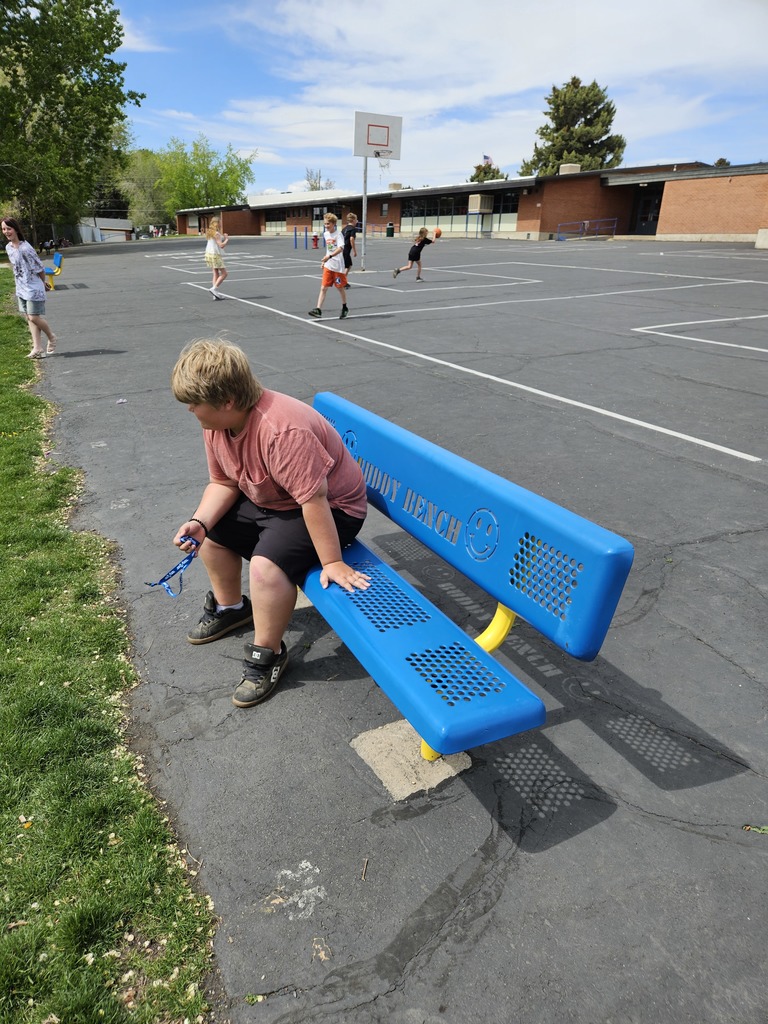 A young boy sits alone on a bright blue metal bench at a school playground. He is looking down and holding a blue lanyard. Other students are playing games in the background. The back of the bench is visible, featuring a friendly smiley face and text (partially obscured) that reads, "BUDDY BENCH".