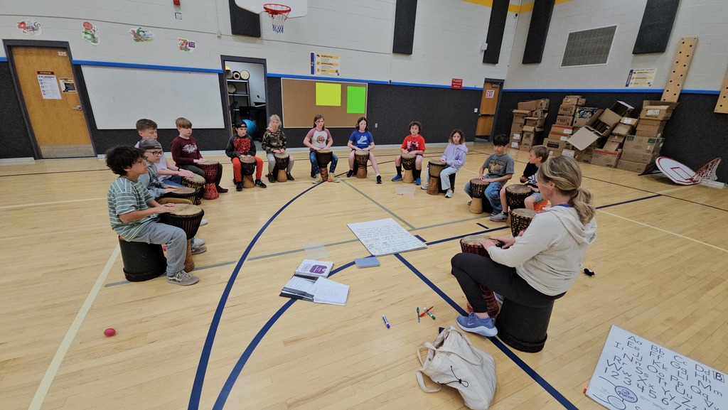 A group of elementary school students practice with hand drums in a school gym, guided by a female instructor. The students are arranged in a large crescent, each seated on a black bucket "drum stool." They are all looking towards the leader, focused on learning a new rhythm. The bright light and polished gym floor give the room an energetic feel.