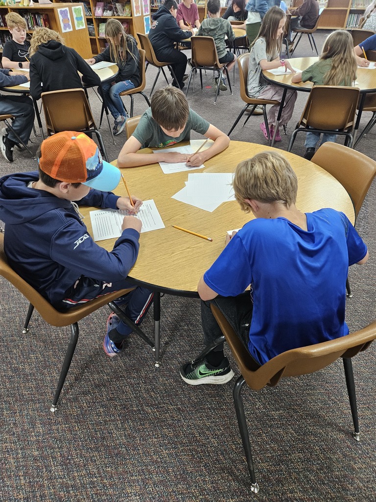  A high-angle shot taken in a school library with patterned grey carpet. A small group of three upper-elementary aged boys are seated at a round wooden table, focused on writing with pencils on lined worksheets. The student on the left wears a blue hoodie and an orange and blue baseball cap. Several other round tables with students working are visible in the background, out of focus.