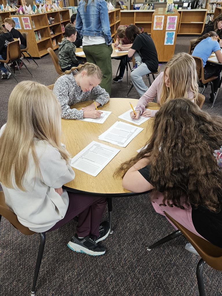 A photograph taken in the same school library environment. A group of four students are gathered around a round wooden table. Two are leaning forward, actively writing on lined paper with pencils, while the other two observe and focus. The student in the foreground on the left has long blonde hair. A teacher or librarian stands partially visible in the background between tables. Shelves filled with books are visible in the background.