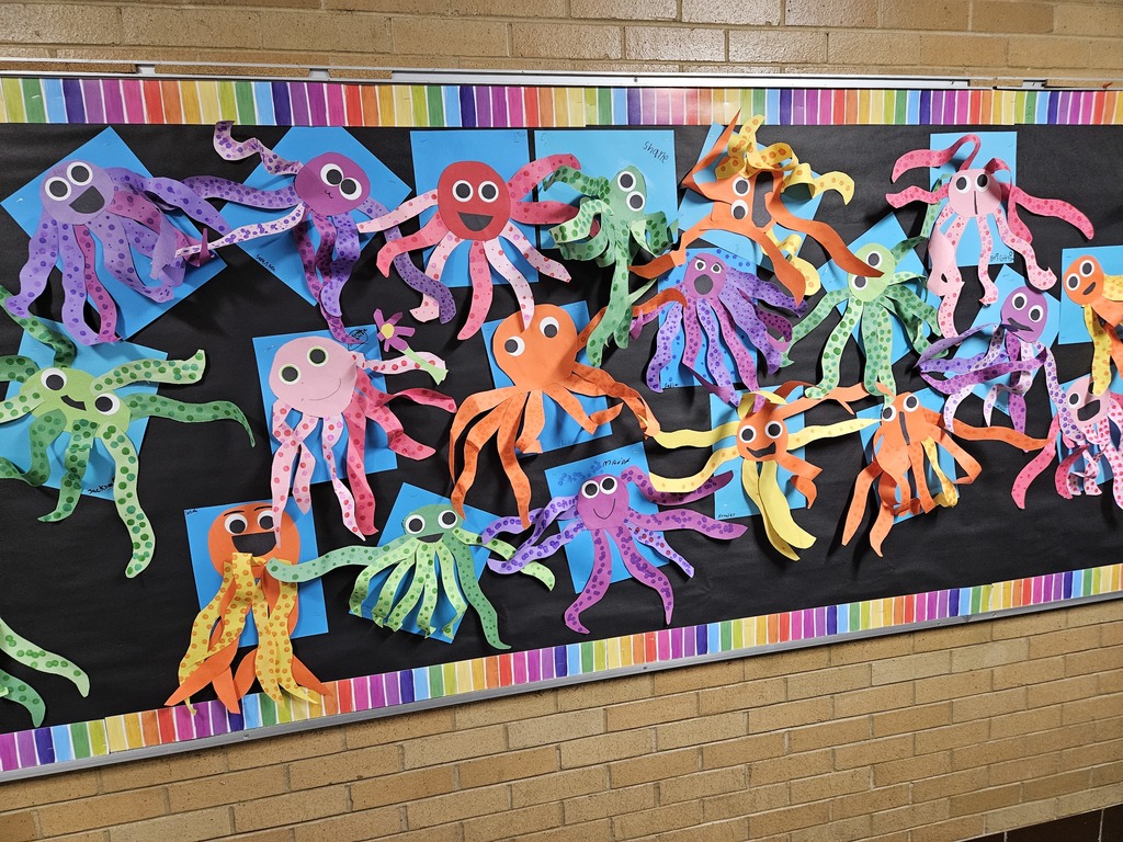 A school bulletin board with a black background and a rainbow-striped border. The board is covered in student-made art projects featuring colorful paper octopuses in shades of purple, green, orange, red, and pink. Each octopus has large googly eyes and long, curly paper tentacles decorated with dots. Some octopuses are mounted on blue square paper backings, creating a vibrant, multi-layered display on a brick hallway wall.
