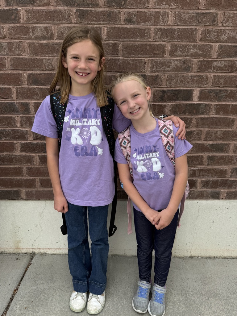Two girl students stand in front of a brick building both wearing purple shirts that say "in my miitary kid  era."