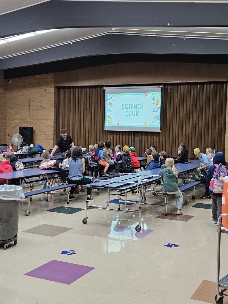 A photograph taken from the back of the Lomond View Elementary school cafeteria showing a "Science Club" meeting in progress. Multiple long, folded-down cafeteria tables with benches are filled with elementary-aged children. Near the middle, a teacher or presenter is standing and talking to a small group. Behind them, a pull-down projection screen displays a slide with a light blue background and the teal text "SCIENCE CLUB," surrounded by simple graphics of magnifying glasses, microscopes, beakers, and test tubes. The room has a distinctive brick-patterned far wall, brown vertical slat curtains behind the screen, and a geometric-tiled ceiling. The floor features colored square patterns and blue "Lomond View Elementary" paw print decals.