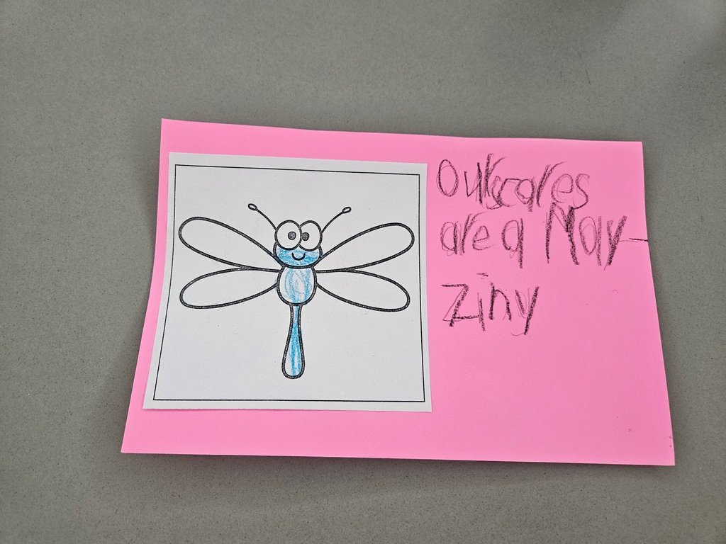 A hand-colored drawing of a blue dragonfly on a bright pink background. Handwritten text reads: "Our scores are a-May-Zing."