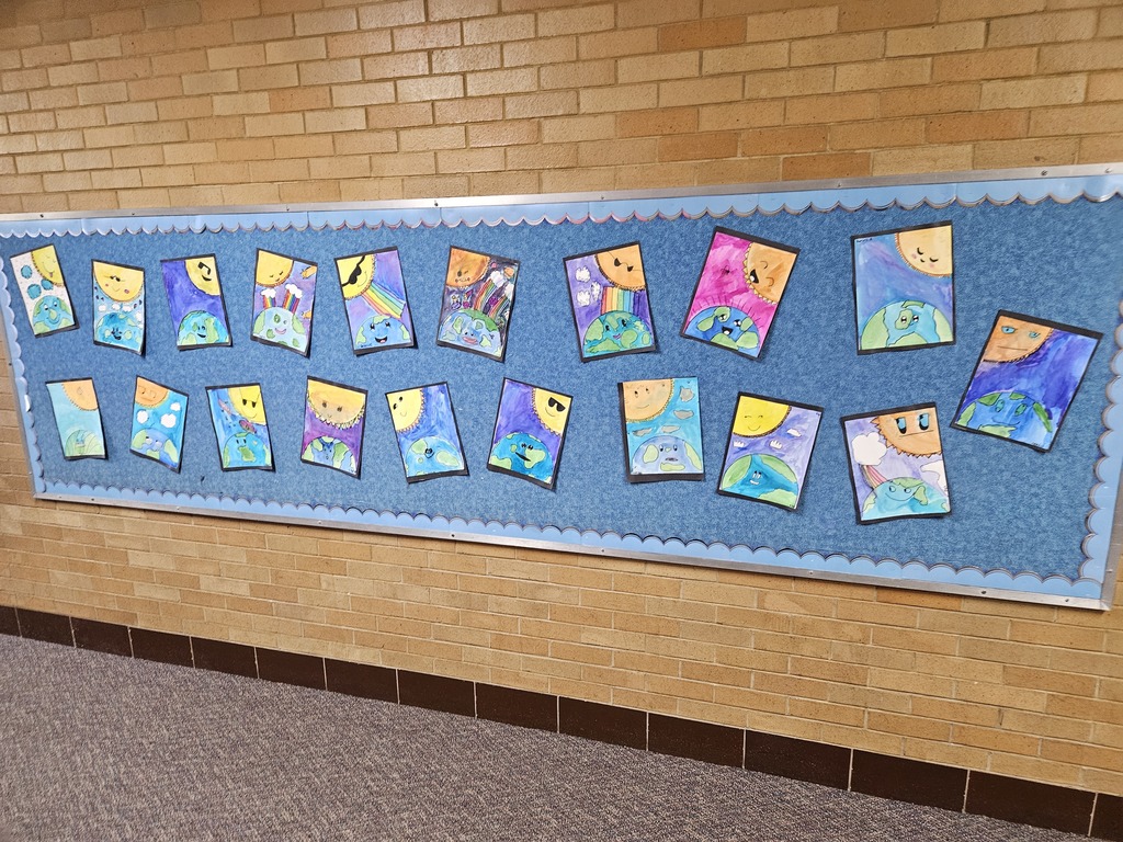 A long hallway bulletin board at Lomond View Elementary with a blue background and scalloped borders. The board is covered in student watercolor paintings celebrating Earth Day. Each painting features a smiling planet Earth looking up at a cheerful sun, many including rainbows, clouds, and vibrant blue or purple skies.