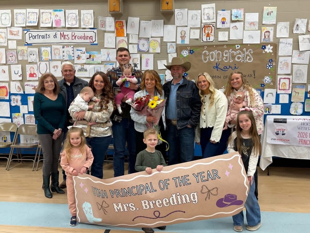 A large group of adults and children pose for a celebratory photo in front of a wall covered in student drawings titled "Portraits of Mrs. Breeding." In the foreground, two children hold a large banner that reads "Utah Principal of the Year Mrs. Breeding." The woman in the center holds a large bouquet of flowers, surrounded by family members and colleagues, including several people holding infants and a man in a cowboy hat. A sign in the background reads "Congrats Kari Admin of the Year!"