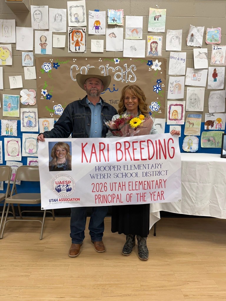 A man in a cowboy hat and a woman holding a bouquet of flowers stand together holding a large celebratory banner. The banner features a headshot of the woman and reads, "Kari Breeding, Hooper Elementary, Weber School District, 2026 Utah Elementary Principal of the Year," along with the Utah Association of Elementary School Principals logo. They are posed in front of a wall filled with hand-drawn student portraits and a sign that says "Congrats Kari."