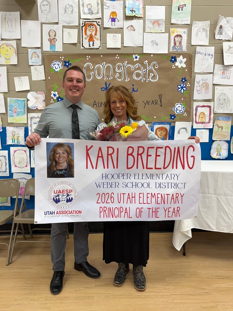 A man in a tie and a woman holding a bouquet of flowers stand together, holding a large celebratory banner. The banner features a headshot of the woman and reads, "Kari Breeding, Hooper Elementary, Weber School District, 2026 Utah Elementary Principal of the Year," along with the Utah Association of Elementary School Principals logo. They are posed in a school setting in front of a wall decorated with hand-drawn student portraits and a "Congrats Kari" sign.
