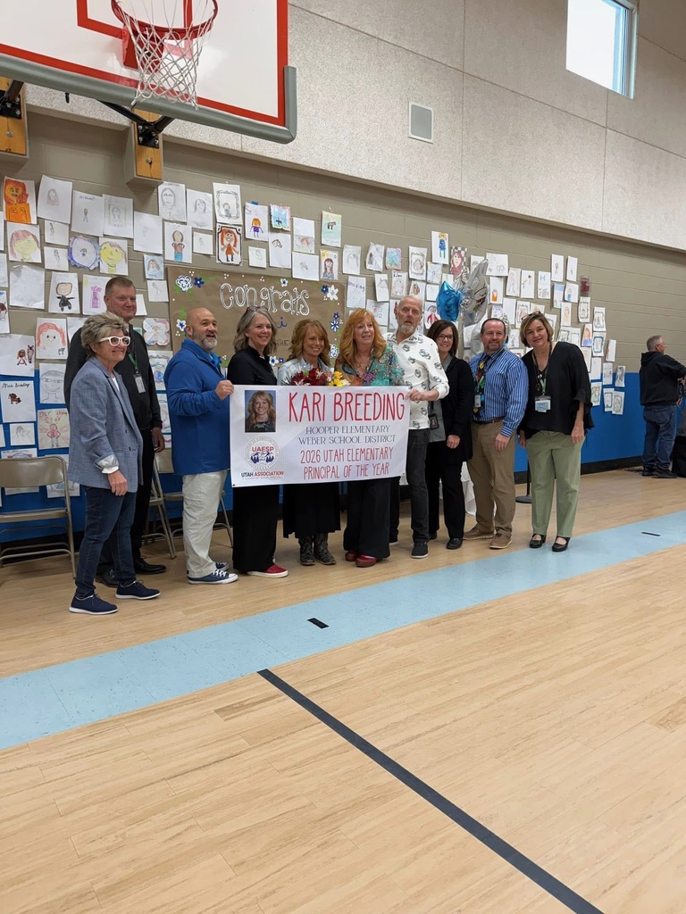 A group of about ten adults stands in a gymnasium, posing together and holding a large celebratory banner. The woman in the center holds a bouquet of flowers. The banner reads, "Kari Breeding, Hooper Elementary, Weber School District, 2026 Utah Elementary Principal of the Year." They are positioned in front of a wall decorated with many student-drawn portraits and a "Congrats Kari" sign, situated directly beneath a basketball hoop.