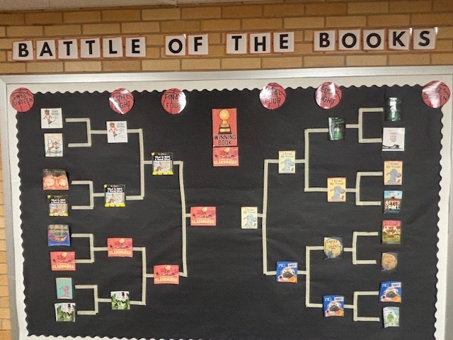 A classroom bulletin board titled "BATTLE OF THE BOOKS" in black and white block letters. The board features a large tournament-style bracket on a black background, decorated with small red basketballs labeled "Sweet Sixteen," "Elite Eight," "Final Four," and "Winning Book." Miniature book covers are placed throughout the bracket to show which books have advanced. In the center, the final matchup is shown between the books "Classmates" and "I Broke My Trunk," with a gold trophy icon at the top representing the winner.