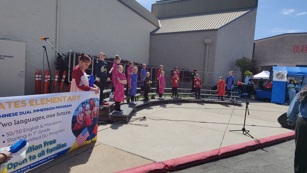 An outdoor performance at Bates Elementary featuring a group of students on a curved stage with choral risers. The students are dressed in various colorful traditional Chinese outfits, including qipaos and tunics in shades of pink, red, and blue. In the foreground, a large banner for the "Chinese Dual Immersion Program" is visible, featuring the slogan "Two languages, one future." A student in a red and white outfit stands next to the banner. The performance is taking place on a sunny day in a paved area next to a school building, with microphones set up on stands.